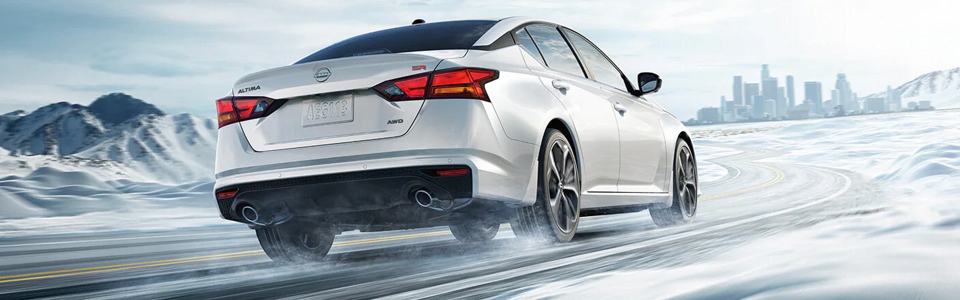 A white Nissan Altima sedan driving on a snowy, icy road, with snow-covered mountains on the left and a city skyline in the distance on the right.